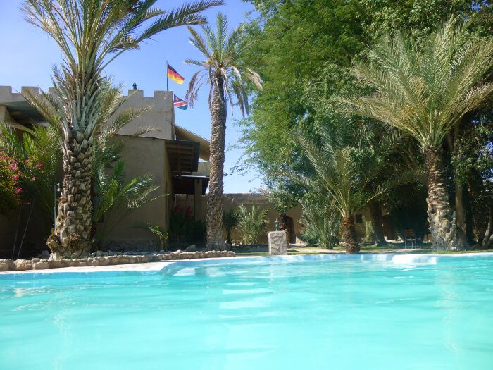 A serene pool at Fort Sesfontein with the tower of the old German fort in the background, flanked by the German and Namibian flags under a clear blue sky.