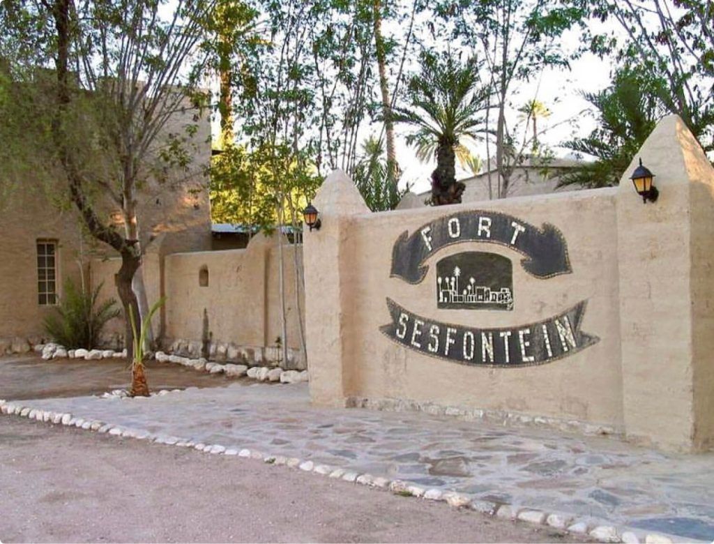 Entrance of Fort Sesfontein featuring the fort's sign, with the lush garden of Fort Sesfontein Lodge visible in the background.