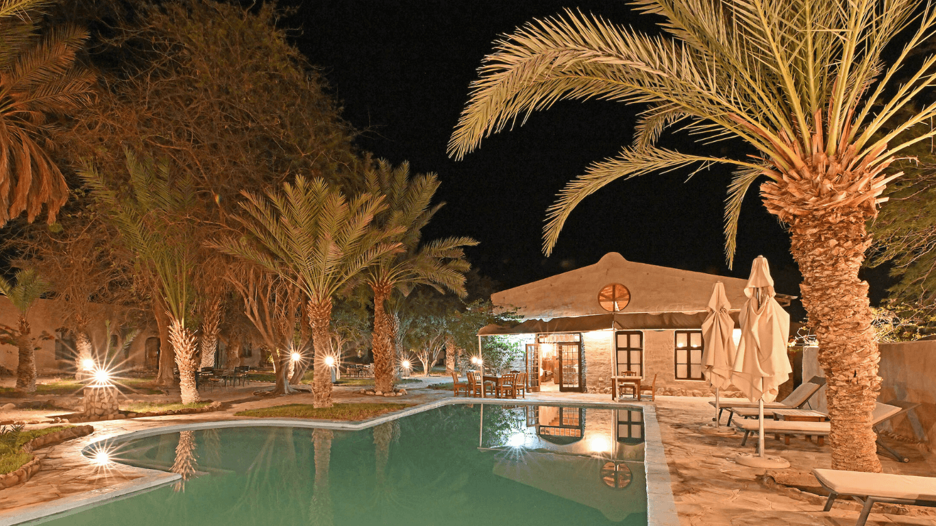 Nighttime view of Fort Sesfontein Lodge's swimming pool with a palm tree to the right, in front of the lodge's restaurant